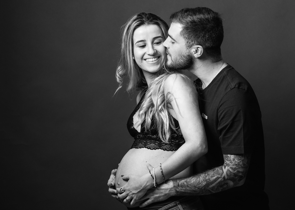 Foto de pareja embarazada en el estudio, foto en blanco y negro con el fondo oscuro, ella lleva un brasier negro de encaje y el lleva una camiseta negra y un gran tatuaje en su brazo, el va a besar a la mama y ambos abrazan la tripa con su bebe