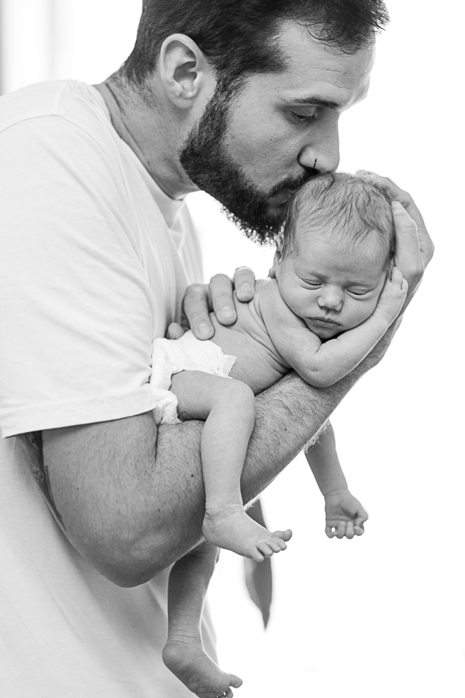 recien nacido newborn durmiendo sobre el brazo de su padre con las piernas colgando mientras su padre le besa tiernamente